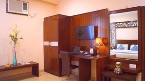 A hotel room with a wooden desk, a chair, a television mounted on the wall, a bed with blue pillows, and a small table with a lamp. - Park Square Hotel And Banquet  (Moradabad)