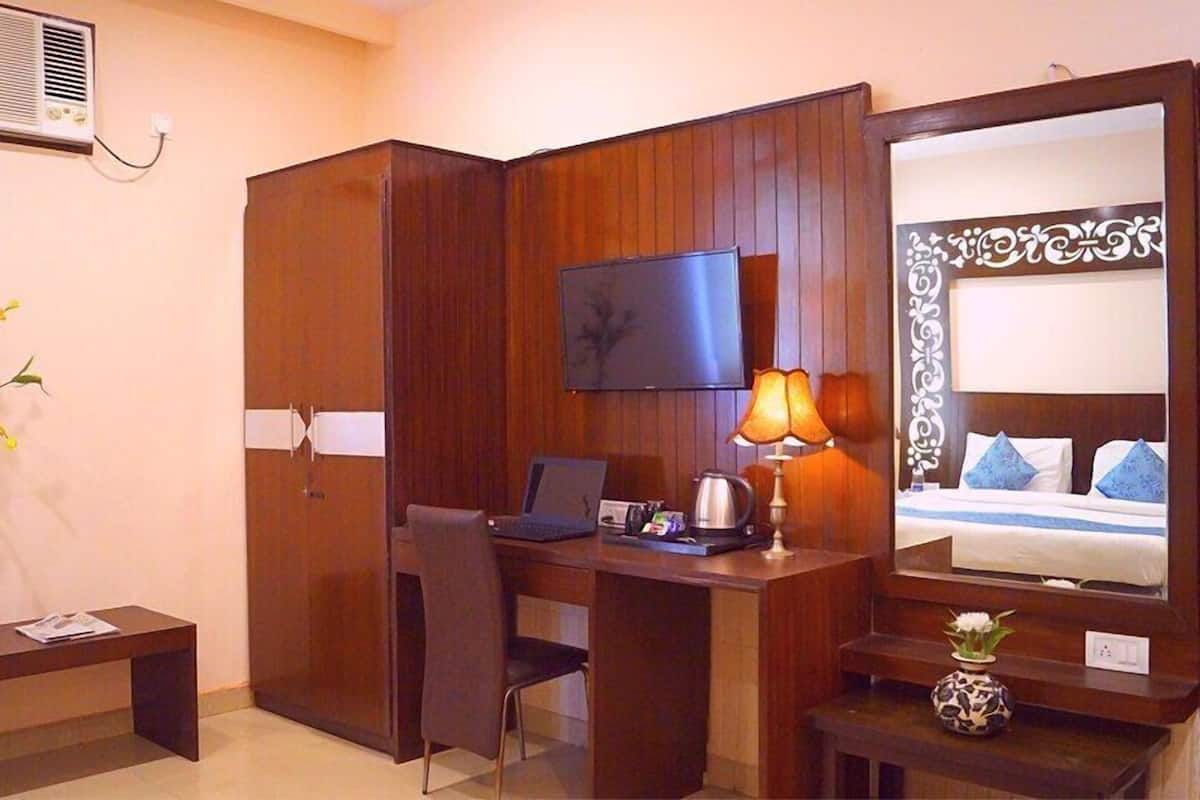 A hotel room with a wooden desk, a chair, a television mounted on the wall, a bed with blue pillows, and a small table with a lamp.