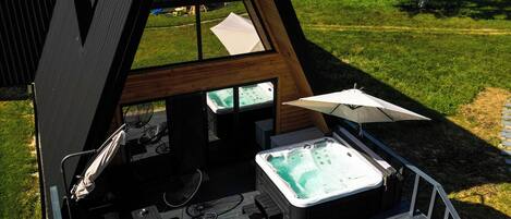 A modern A-frame structure with a hot tub, outdoor seating, and a view of the countryside.
