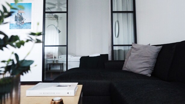 Ein modernes Schlafzimmer mit einer schwarzen Couch, einem Nachttisch mit einem Buch und einem großen Fenster mit Vorhängen.
