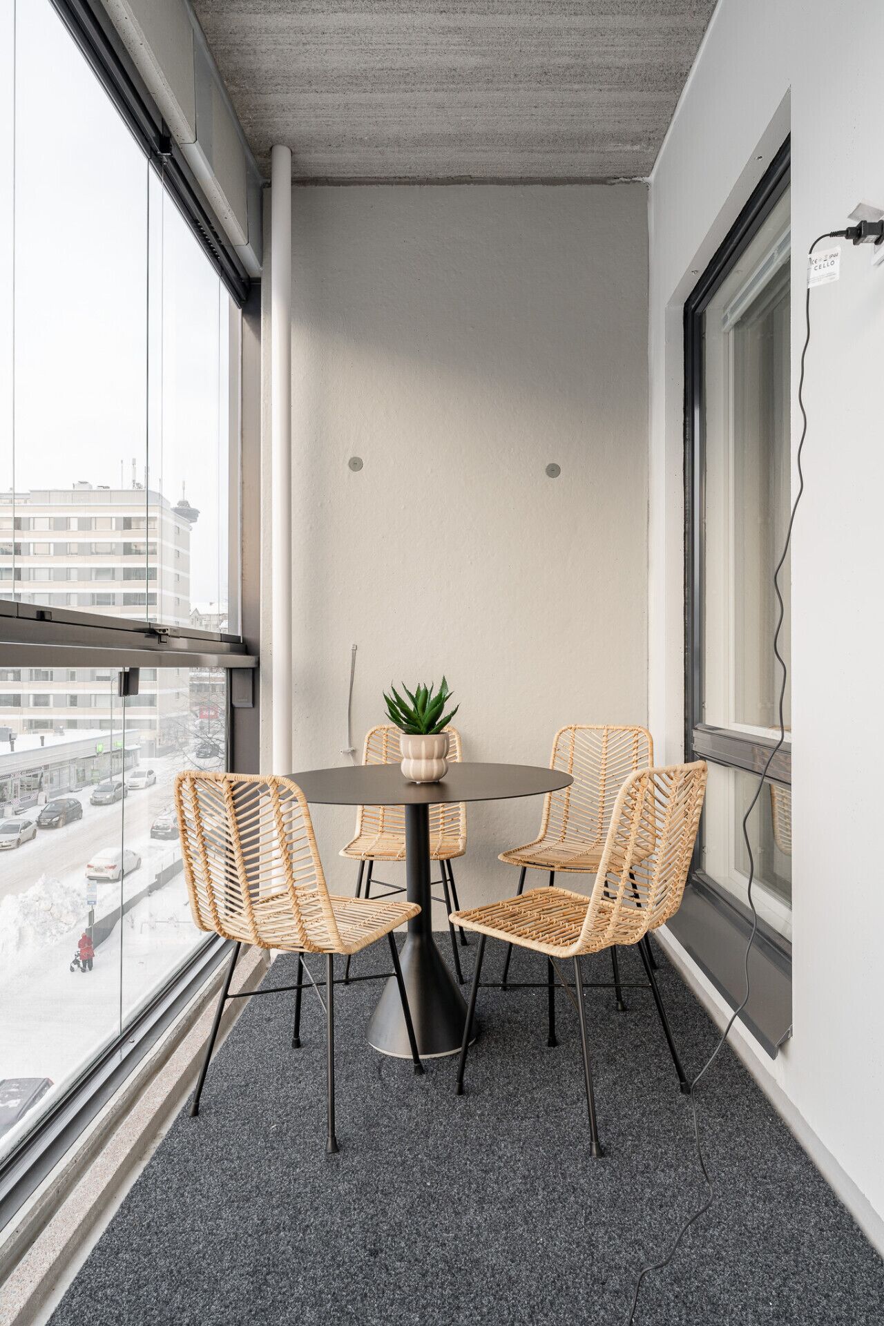 Moderni huone, jossa on pyöreä pöytä, neljä punottua tuolia, ruukkukasvi ja suuri ikkuna, josta avautuu näkymä lumiseen kaduun.