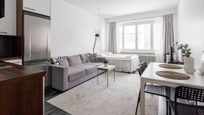 A modern, well-lit living room with a grey sofa, a dining table with chairs, a kitchen area with a fridge, and a large window providing natural light.