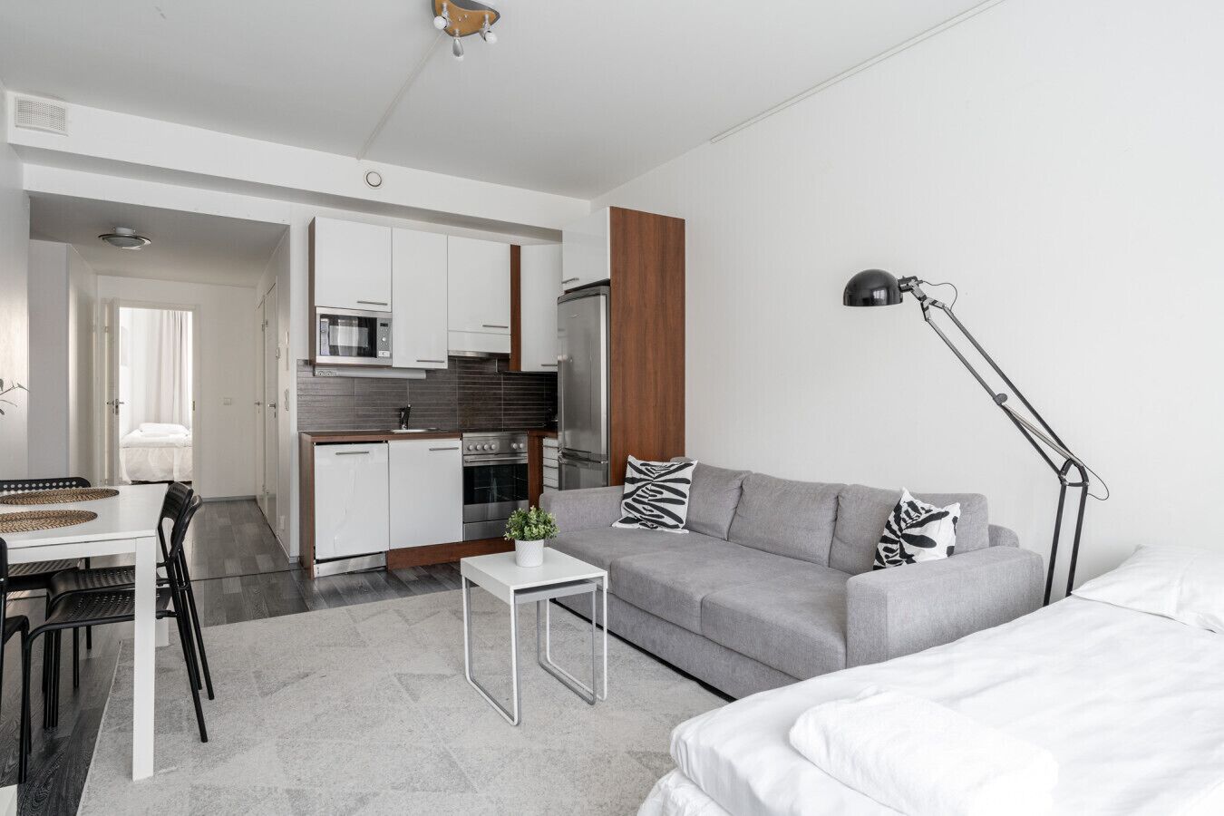 Ein moderner, gut beleuchteter Raum mit grauem Sofa, einem kleinen weißen Tisch, einem Essbereich mit Stühlen, einer Küchenzeile und einer Nachttischlampe.