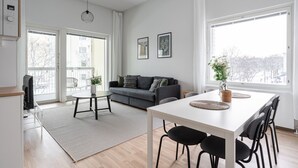 A modern living room with a grey sofa, a white dining table with black chairs, and a large window providing a view of the outdoors.