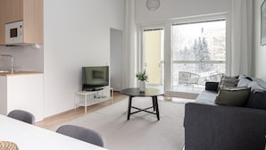 A modern living room with a grey sofa, a small round table with a plant, a TV on a white stand, and a balcony with a view of trees.
