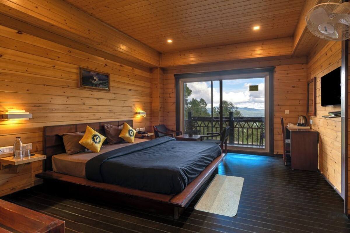 A wooden cabin bedroom with a bed, bedside tables, a TV, and a balcony with a view.