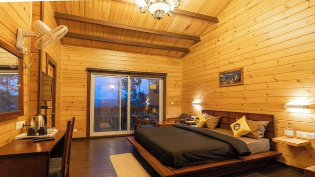 A wooden cabin bedroom with a bed, bedside table, lamp, and a view through a sliding glass door. - The Hosteller Kasar Devi (Almora)