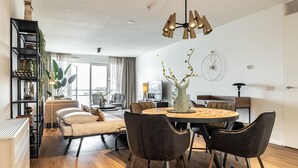 A modern living room with a round dining table, dark chairs, and a chandelier. There is a bookshelf with plants and a sofa with cushions.