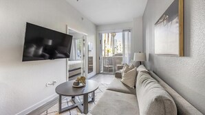 Een moderne woonkamer met een flatscreen televisie aan de muur, een bank, een klein rond tafeltje met een decoratief object, en een groot raam met uitzicht op het omliggende landschap.