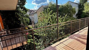 A balcony with tiled flooring and a metal railing, overlooking a garden with various plants and a building in the background.