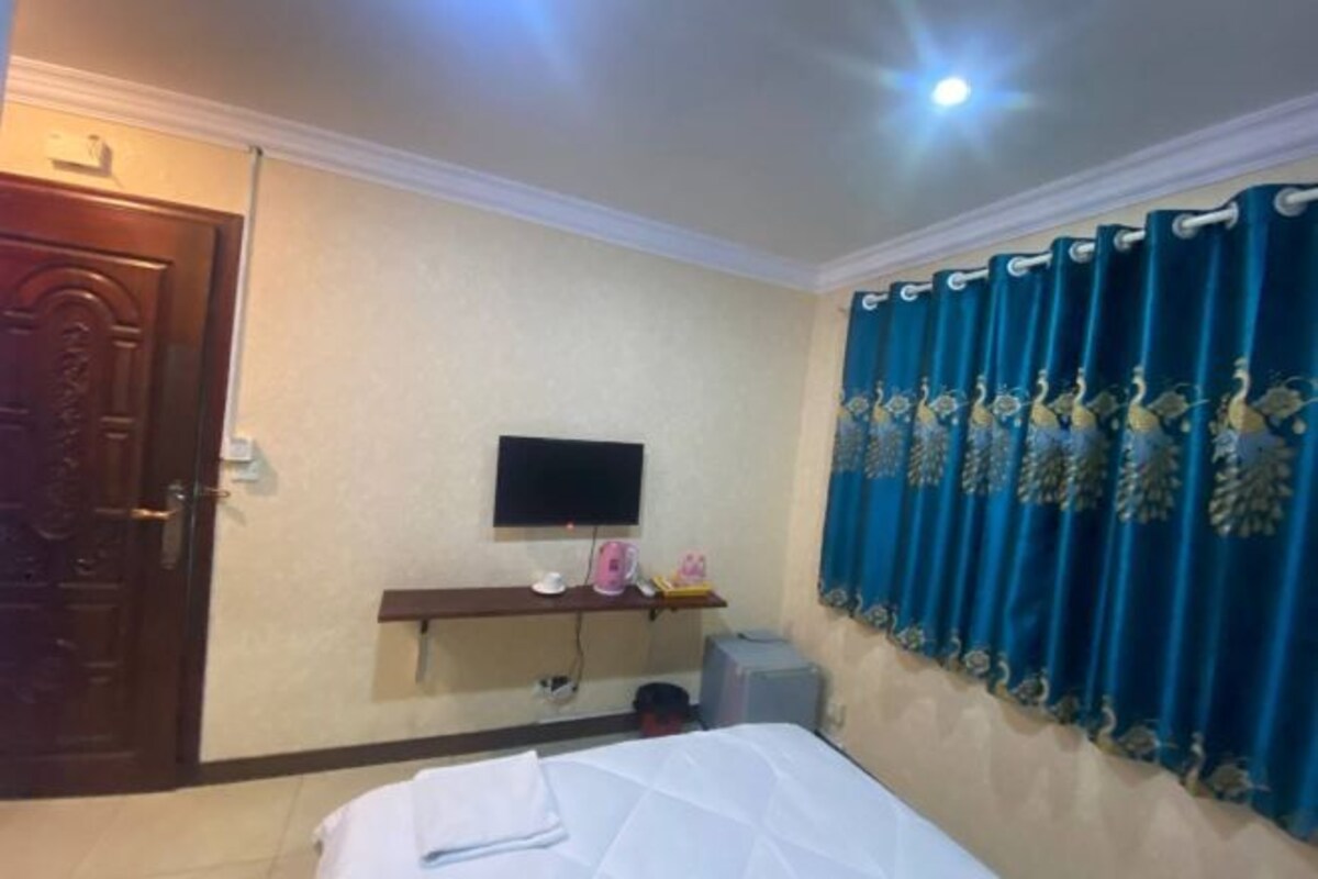 A hotel room with a bed, a television mounted on the wall, a shelf with items, and a curtain.