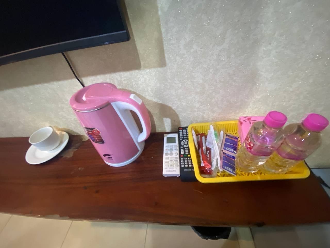A wooden table with a pink mug, a white cup, a calculator, a yellow tray with water bottles and snacks, and a TV mounted on the wall.