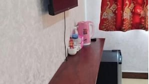 A room with a TV mounted on the wall, a small wooden shelf with a kettle and cups, and a red curtain with a peacock design. - TWILIGHT INN (Phnom Penh)