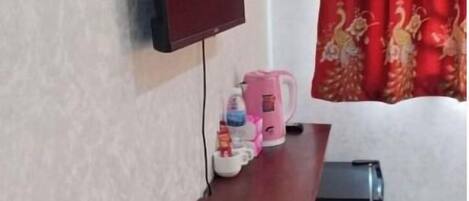 A room with a TV mounted on the wall, a small wooden shelf with a kettle and cups, and a red curtain with a peacock design.