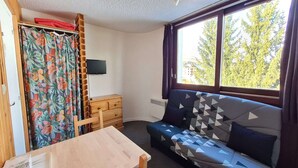 A room with a wooden table, a sofa with geometric patterned cushions, a wooden dresser, a floral curtain, and a window with a view of trees.