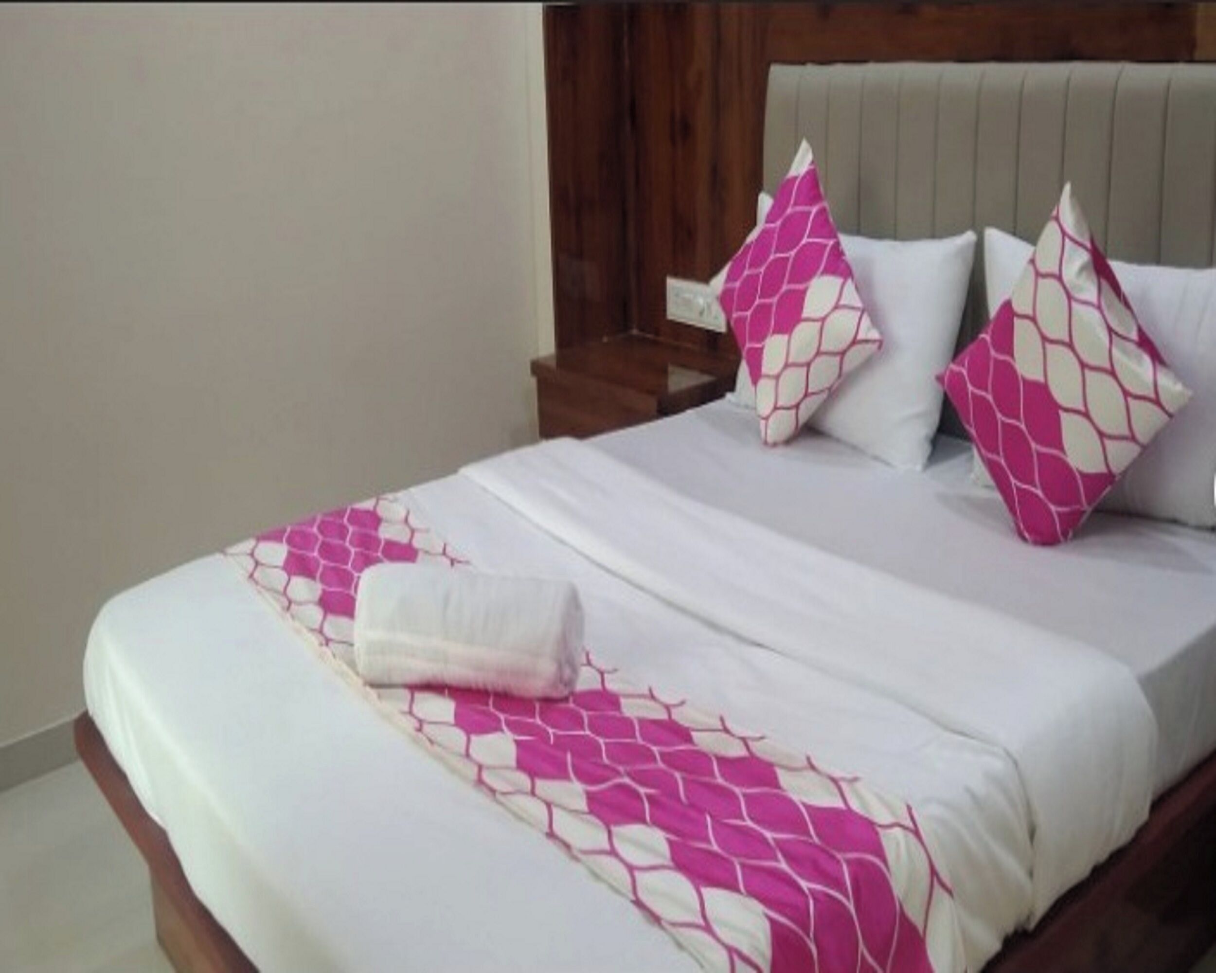 Een net opgemaakt bed met een deken in wit en roze dessin, twee bijpassende kussens en een houten hoofdbord.