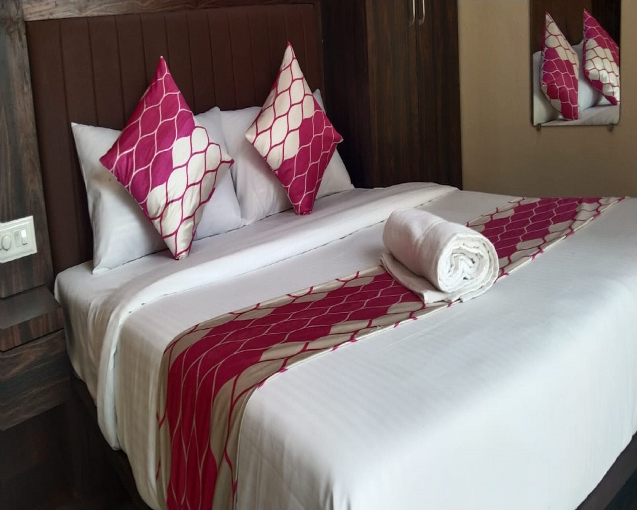 A neatly made bed with white bedding and pink patterned pillows, a rolled white towel, and a wooden headboard.
