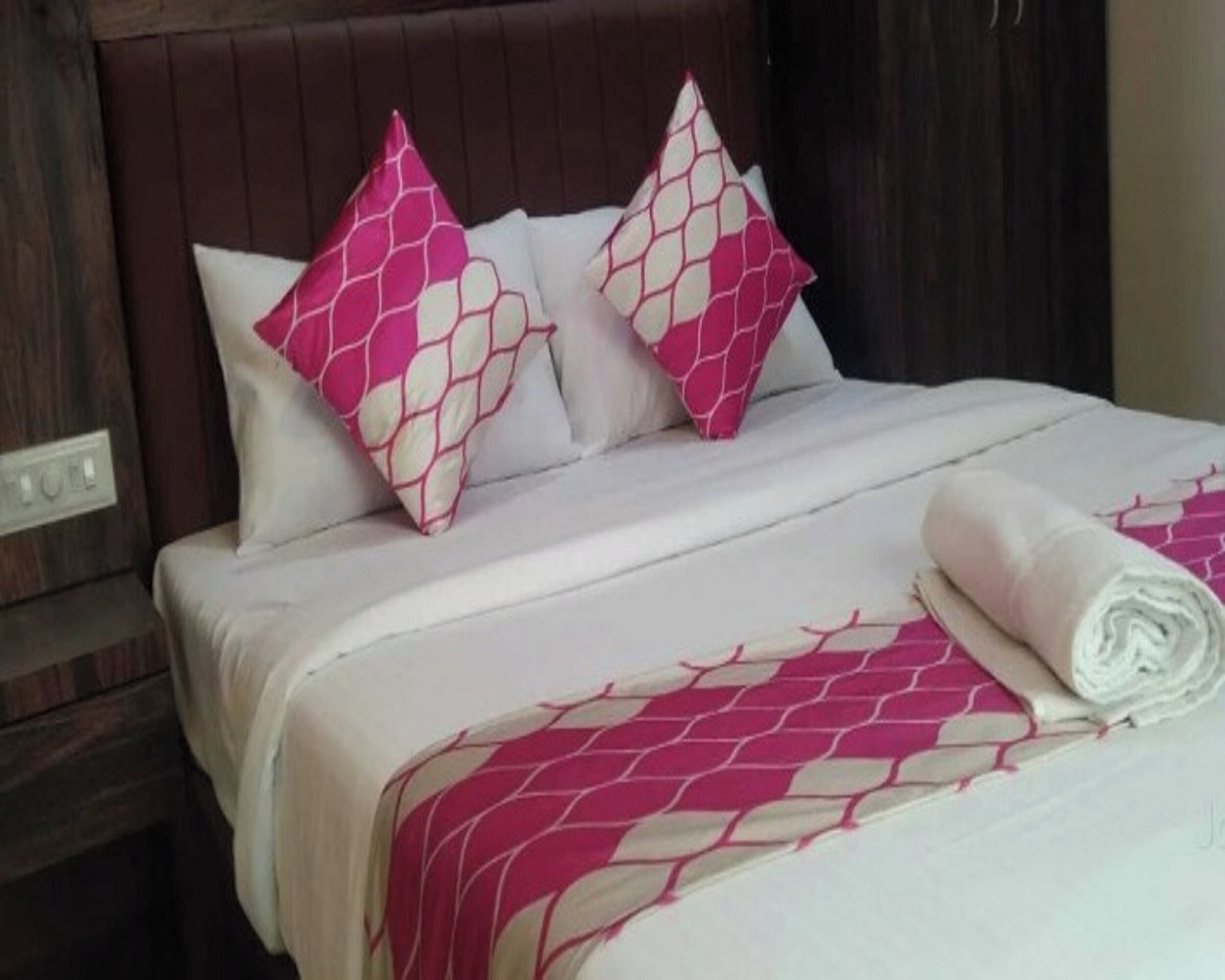 A neatly made bed with a pink and white patterned bedspread and pillows, a wooden headboard, and a power outlet on the wall.