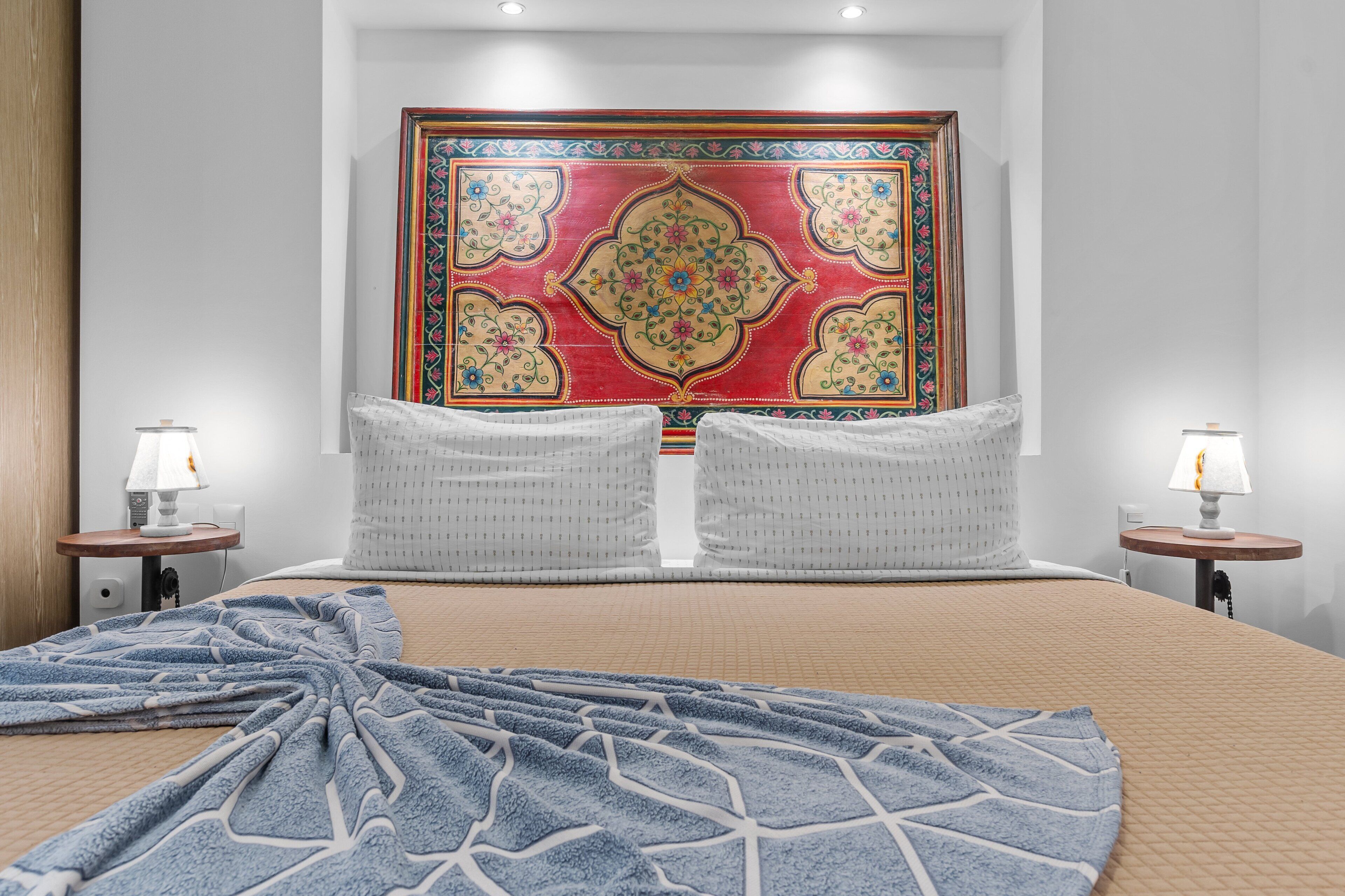 A hotel room with a large bed, two bedside tables with lamps, a decorative headboard, and a patterned throw blanket.