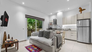 Een moderne woonkamer met een grijze bank, een salontafel en een kleurrijk vloerkleed. In de achtergrond is een keuken zichtbaar met roestvrijstalen apparatuur en houten kastjes.