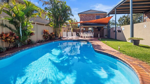 A swimming pool with a brick border, surrounded by a grassy area and a covered patio with chairs. - Romantic King Spa Suite Perfect for Couples in Coffs Harbour (Coffs Harbour)