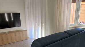 Ein modernes Wohnzimmer mit einem dunkelblauen Sofa, einem Flachbildfernseher auf einem Holzboden und einem großen Fenster mit transparenten Vorhängen.