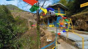 A blue building with yellow accents, a staircase leading to the entrance, and colorful flags hanging from a tree. - The Hosteller Mussoorie By The Streamside, Kempty (Mussoorie)