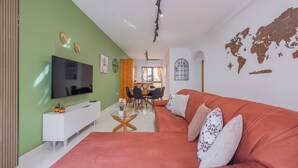 Una sala de estar moderna con un sofá grande color naranja, un mueble blanco para entretenimiento y un televisor de pantalla plana. Hay un área de comedor con una mesa y sillas, y un mapa mundial en la pared.