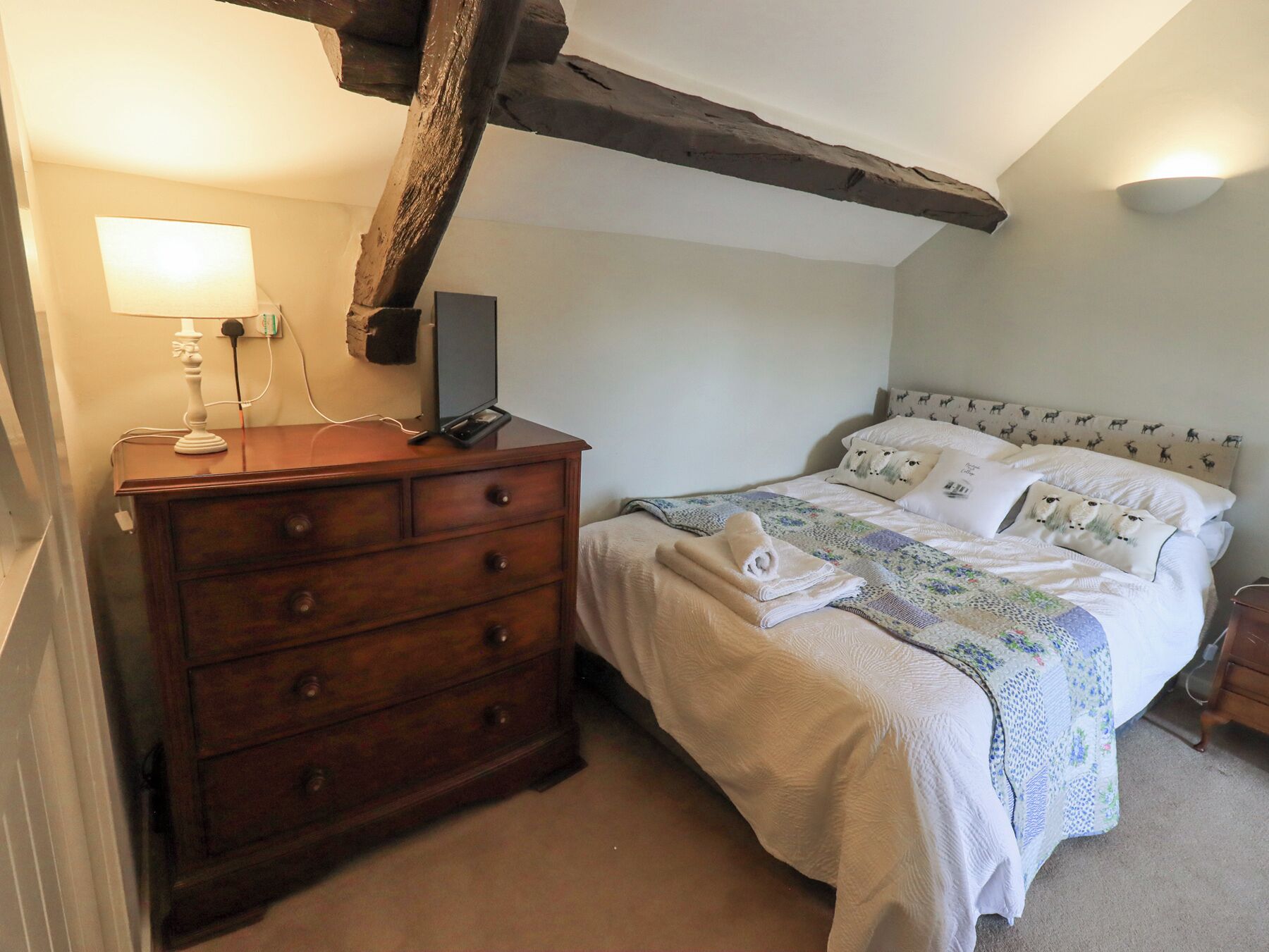 A bedroom with a wooden dresser, a bed with patterned bedding, and exposed wooden beams on the ceiling.