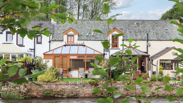 A riverside property with a house featuring a conservatory, a stone wall, and a garden. - Beckside Cottage (Penrith)