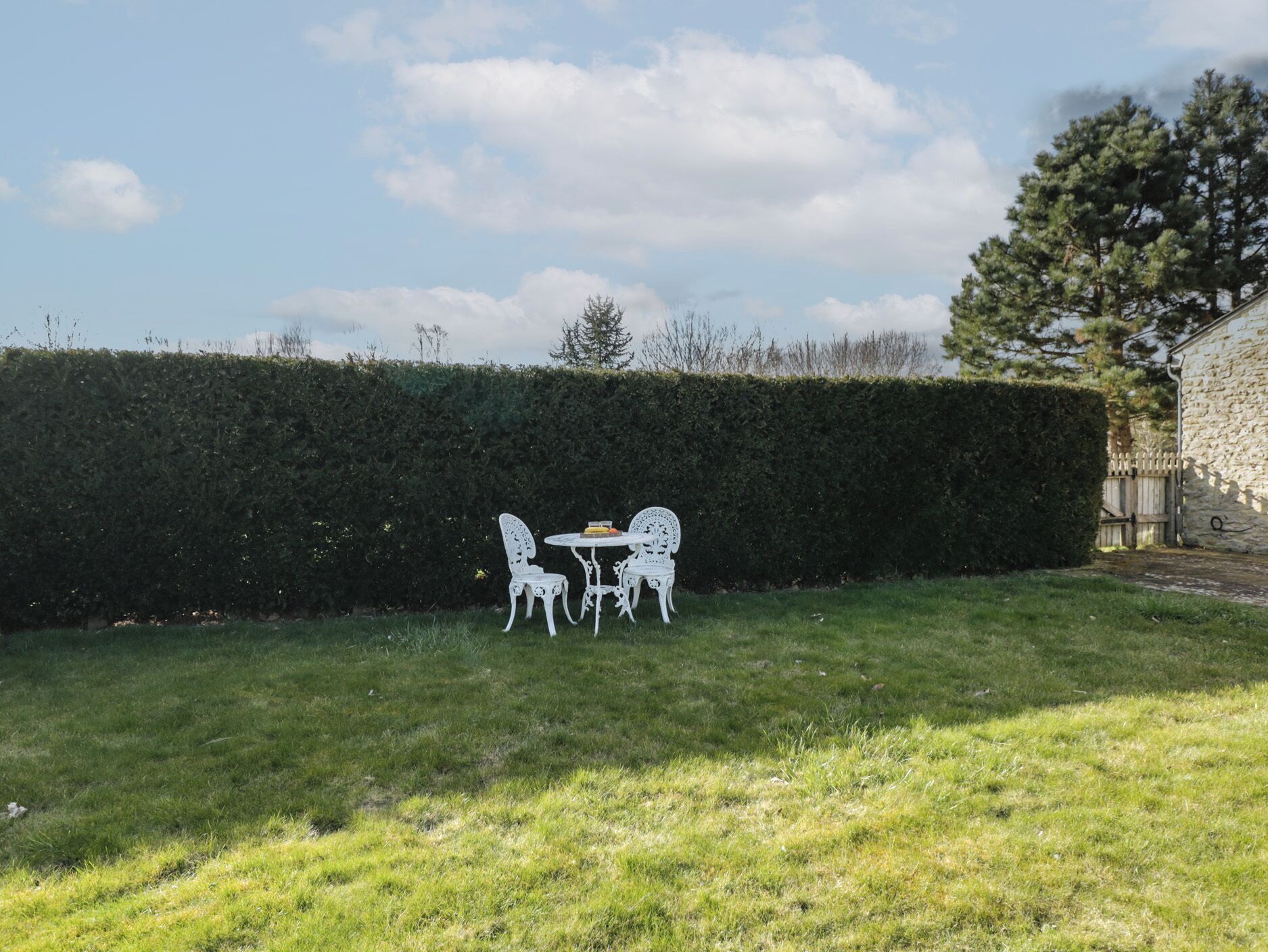 Een tuin met een witte metalen tafel en stoelen, een haag en een stenen gebouw op de achtergrond.