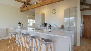A modern kitchen with a large island, bar stools, wooden beams, and a clock on the wall. - South Barn Cottage (Faringdon)