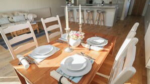 A dining area with a wooden table set for four, white chairs, and a kitchen in the background. - South Barn Cottage (Faringdon)