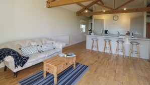 A modern living room with a sofa, coffee table, and a kitchen area with bar stools. - South Barn Cottage (Faringdon)