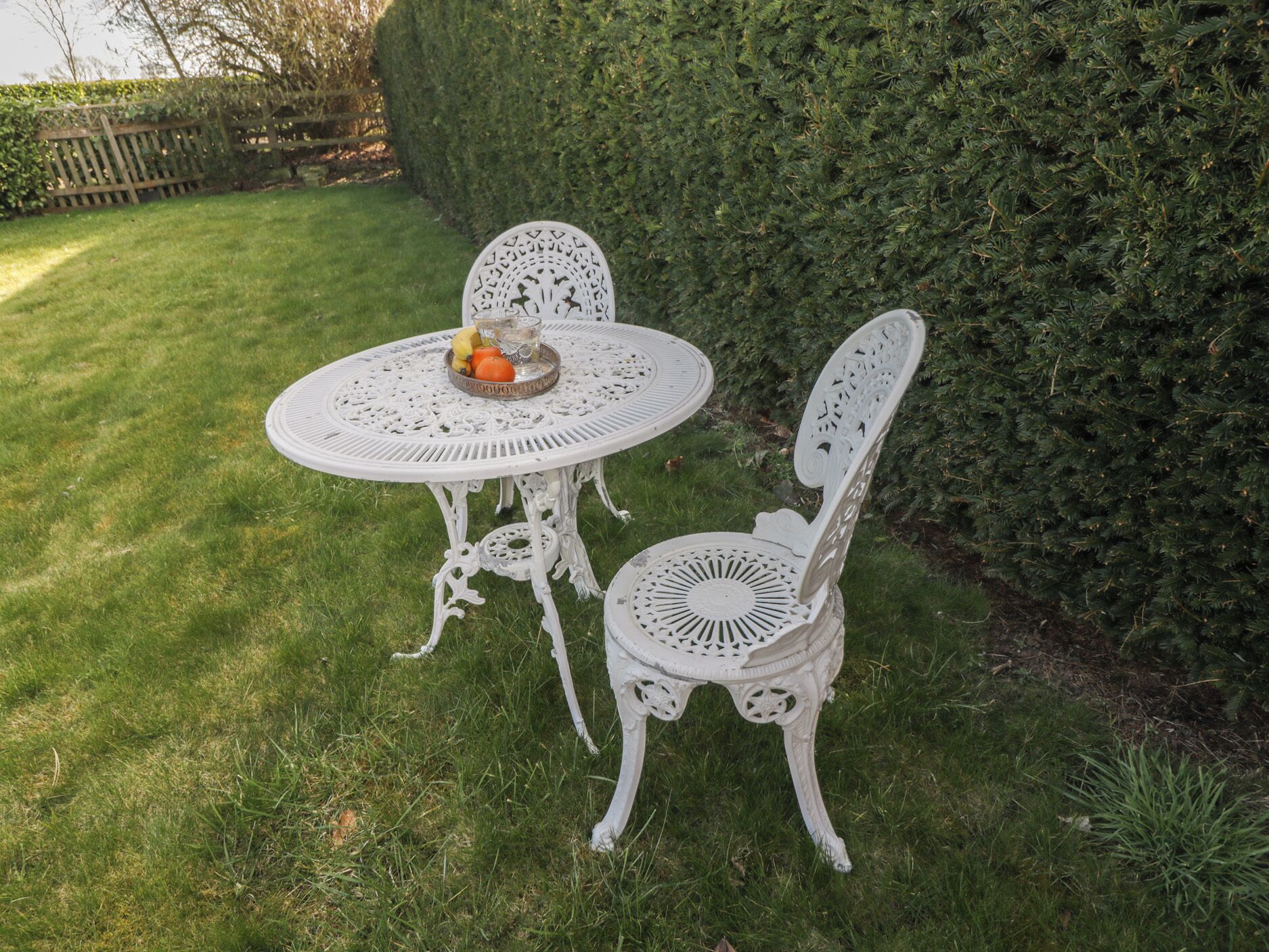 Een tafel en stoelen van wit ijzer op een gazon, met een haag op de achtergrond.