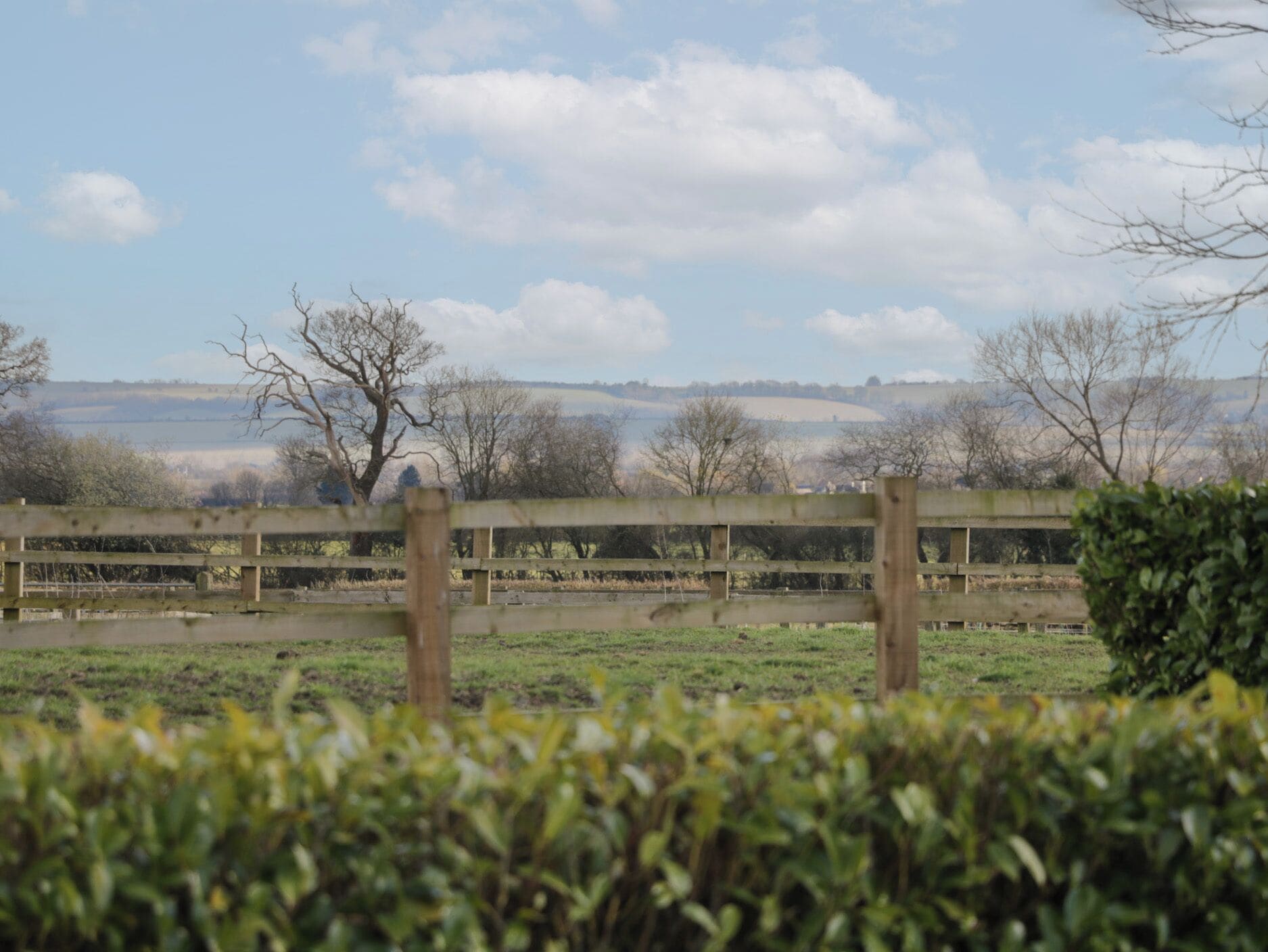A rural landscape with a wooden fence, open fields, and distant hills under a partly cloudy sky.