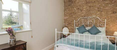 Un dormitorio con una cama de metal blanco, una mesita de noche de madera, una ventana con vistas a árboles y una pared de ladrillo.