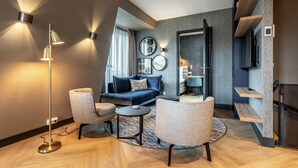 A modern living room with a sofa, two armchairs, a coffee table, and a floor lamp. There are wall-mounted lights and a TV on a shelf.
