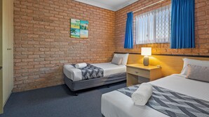 Une chambre d’hôtel avec un mur en briques, deux lits, une table de chevet avec une lampe et une fenêtre avec des rideaux bleus.