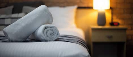 Una cama bien hecha con una toalla enrollada, una lámpara en la mesita de noche y una pared de ladrillo al fondo.