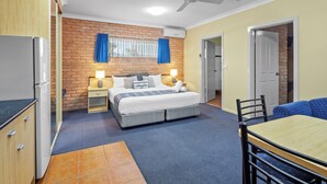 Une chambre à coucher comprenant un lit, des tables de chevet, un ventilateur de plafond, une fenêtre avec des rideaux, un mur en briques et une porte donnant sur une autre pièce.