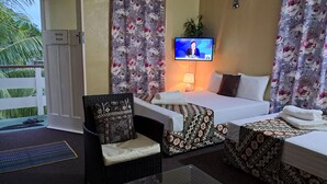 A hotel room with a bed, two bedside tables, a chair, a TV, and a balcony with a view of palm trees.