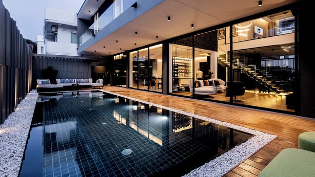 Una casa moderna con piscina, terraza de madera y puertas de cristal que dan a un interior bien iluminado.