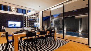 Un comedor moderno con una mesa de madera, sillas negras y un tapete a rayas blanco y negro. Un televisor grande está montado en la pared, y se ve un expositor de zapatos al fondo.
