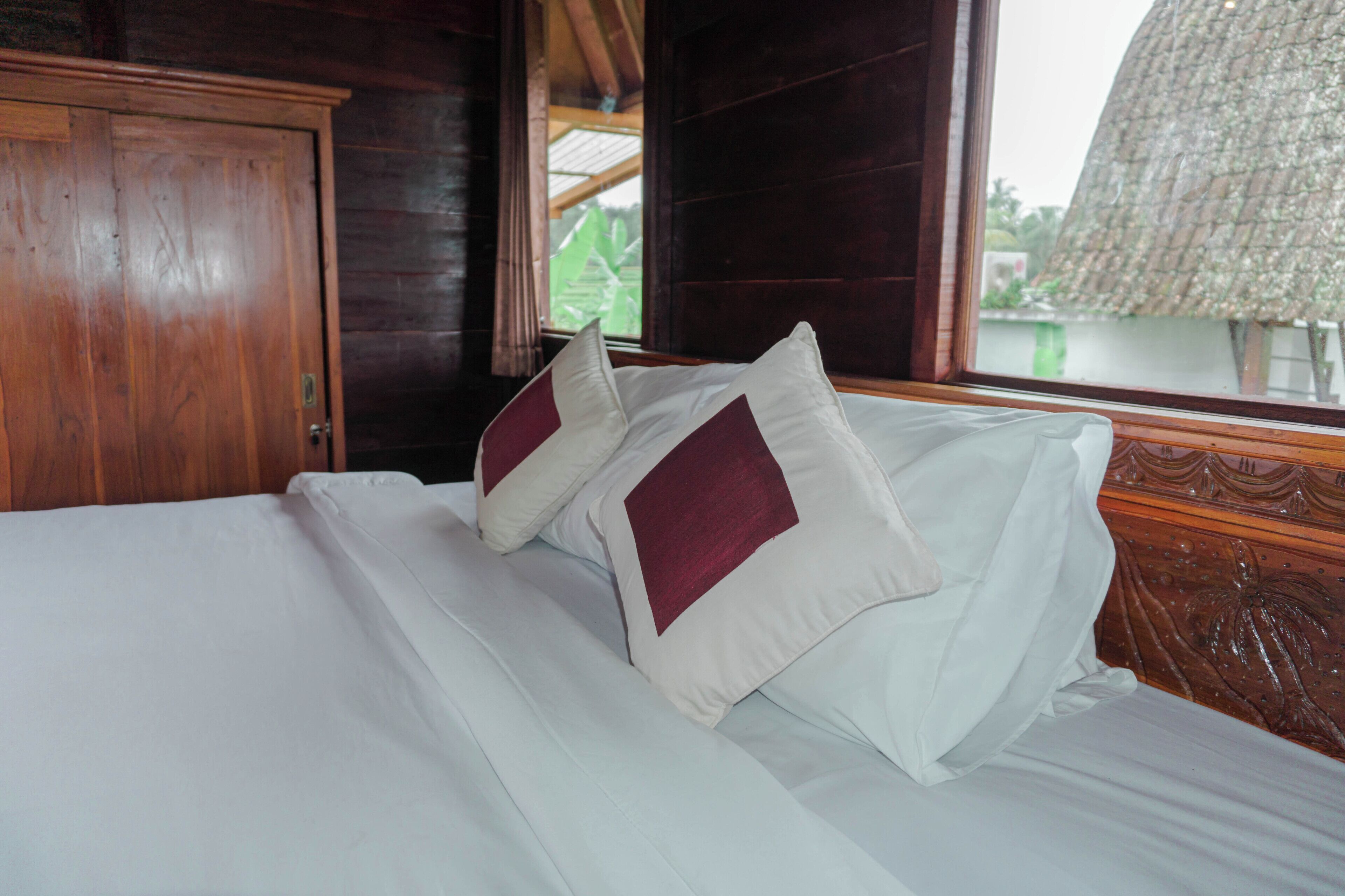 A bed with white linens and two pillows, a wooden wardrobe, and a window with a view of greenery.