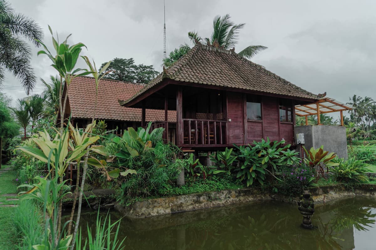 Une maison traditionnelle en bois, avec un toit de tuiles, entourée d’une végétation luxuriante et d’un petit étang.