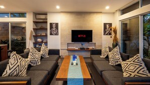 Un salon moderne avec un téléviseur à écran plat fixé à un mur en pierre, deux canapés gris ornés de coussins à motifs, une table basse en bois avec un chemin de table bleu, et une bibliothèque contenant des objets décoratifs.
