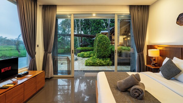 Une chambre d’hôtel moderne dotée d’un grand lit, d’une télévision et offrant une vue sur un jardin et un bâtiment, le tout visible à travers une porte vitrée.