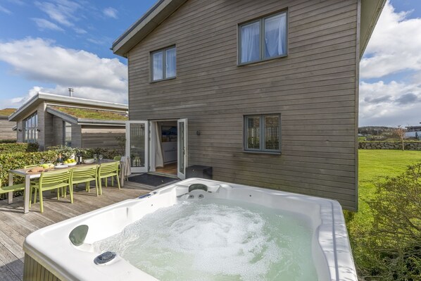 A modern house with a hot tub, a dining area with a table and chairs, and a grassy yard.