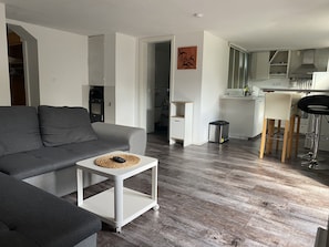 En moderne stue med en grå sofa, et hvidt sofabord og et køkkenområde med en hvid bordplade og barstole.